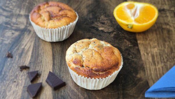 Muffins auf braunem Holztisch mit blauer Serviette, Schokostückchen und Mandarinen.