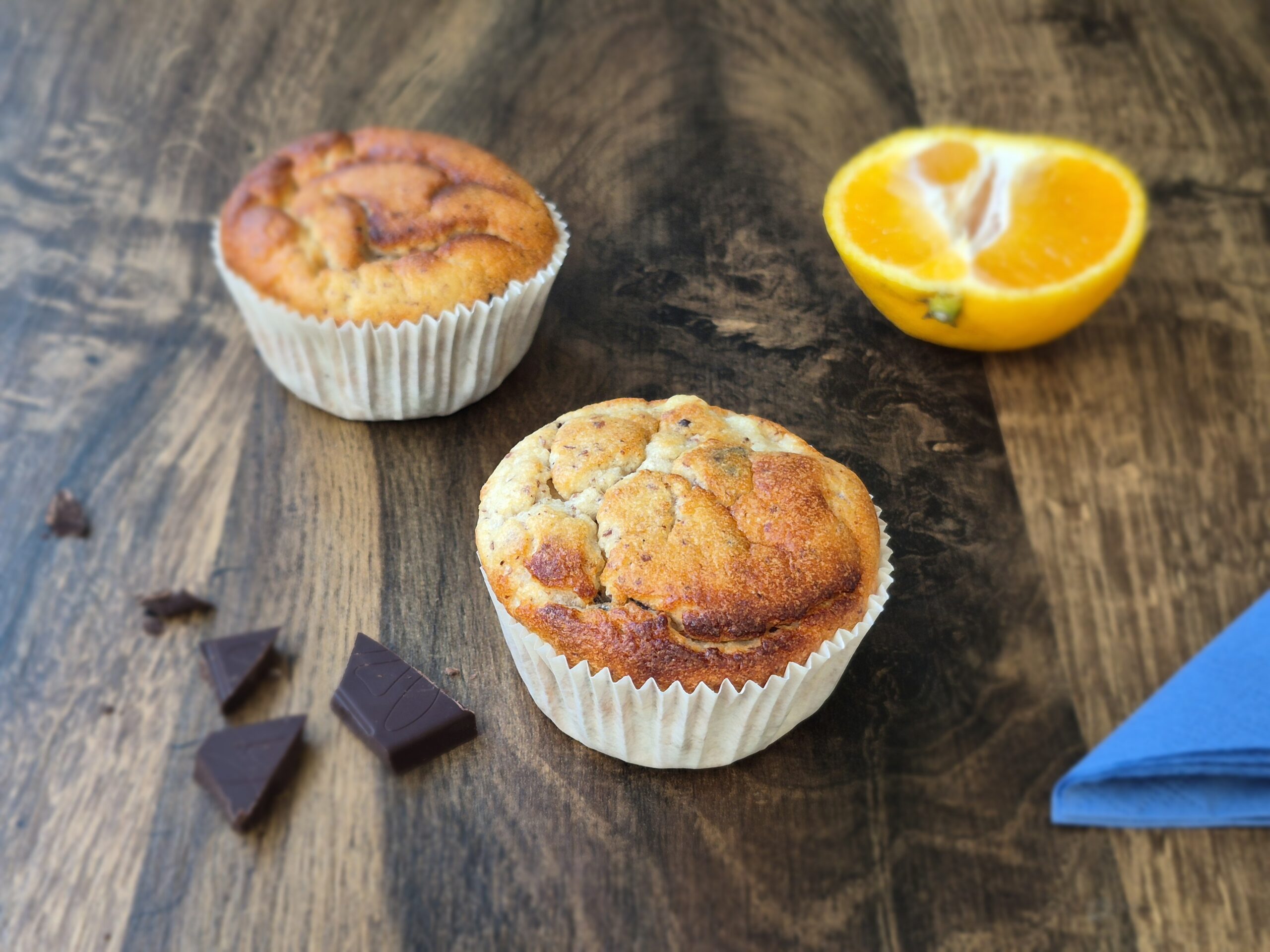 Muffins auf braunem Holztisch mit blauer Serviette, Schokostückchen und Mandarinen.